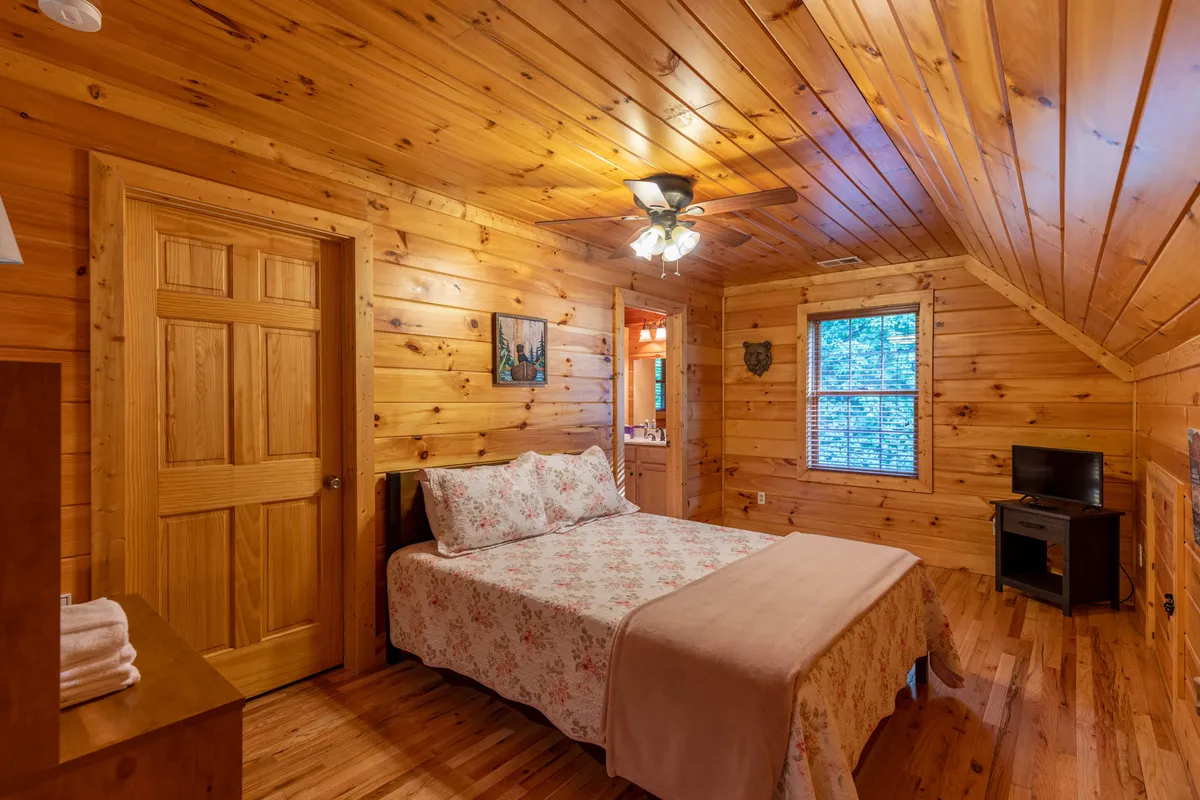 A serene bedroom with a comfortable bed dressed in floral bedding, wooden walls, a ceiling fan, and abundant natural light streaming through the window—an inviting and restful private retreat.