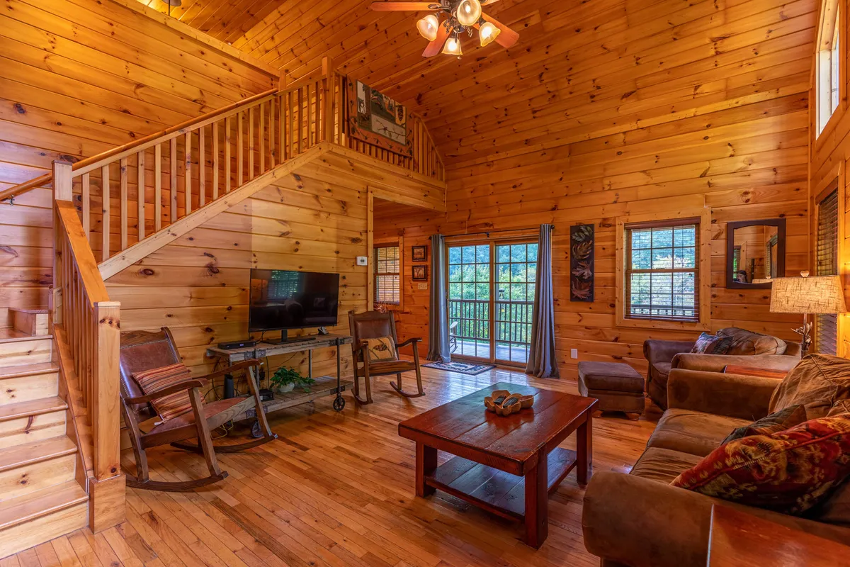 A welcoming living room with a central coffee table, large windows letting in natural light, a comfortable sofa, rocking chair, and a view of the wooden staircase. The space exudes warmth and is designed for guests to relax, entertain, or enjoy intimate gatherings.