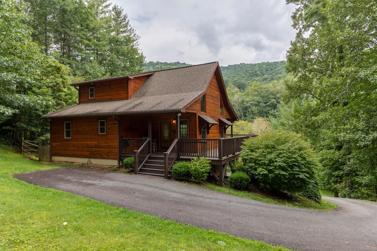 The image showcases the charming wooden exterior of a three-bedroom cabin, nestled among lush trees with a welcoming front porch and a beautifully landscaped yard. The inviting porch is perfect for relaxing, while the serene surroundings hint at endless outdoor adventures and peaceful riverfront moments.