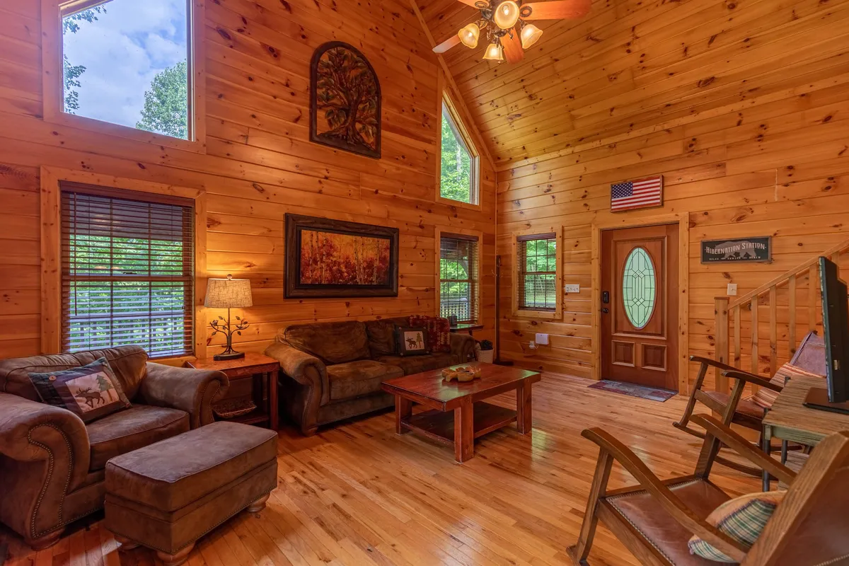 A spacious living room filled with warm wooden tones, large windows that invite natural light, comfortable seating arranged for conversation or relaxation, and artwork on the walls. The setting feels welcoming and ideal for gathering with friends or family, playing games, or simply soaking in the mountain ambiance.