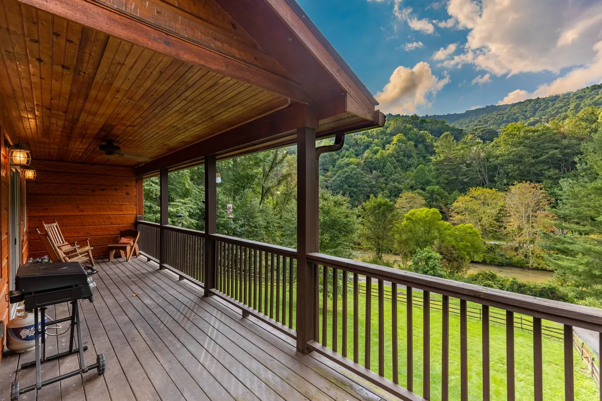 This image showcases an exterior view of a covered wooden porch featuring inviting rocking chairs, a grill, and an expansive view of the surrounding landscape. The open railing draws the eye to the lush outdoors, suggesting proximity to nature and riverfront relaxation.